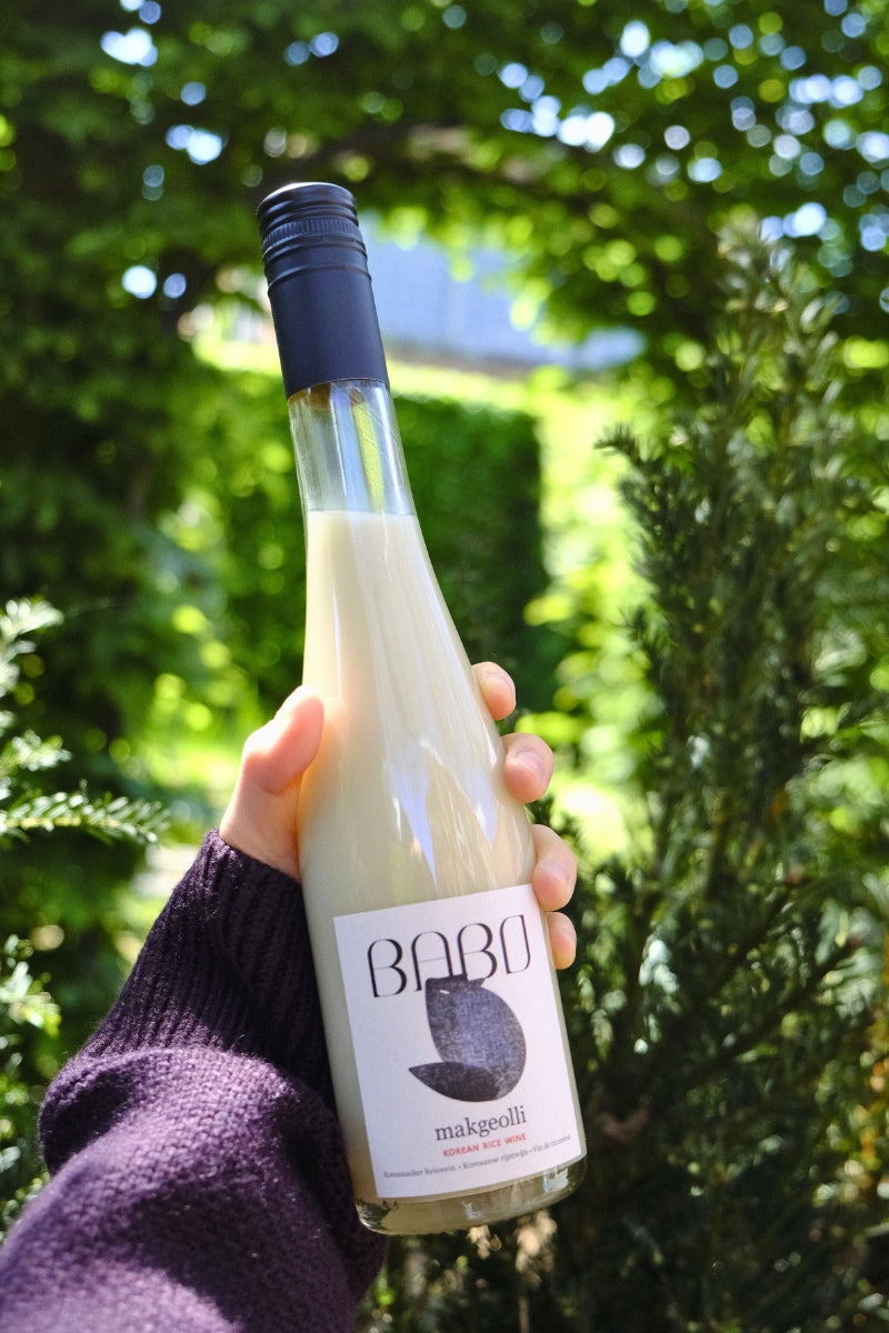 Hand holding up a bottle of Babo Makgeolli outside in sunny weather with greenery background.