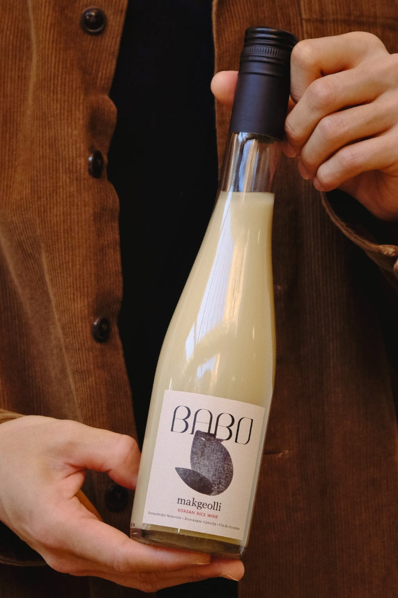 Person presenting a bottle of Babo Makgeolli, artisanal Korean rice wine brewed in Belgium, held with both hands.