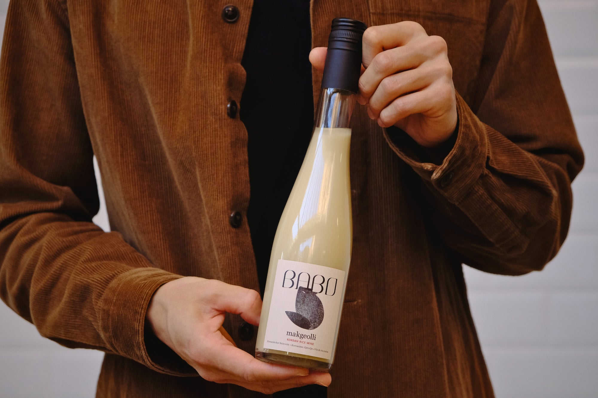 Person presenting a bottle of Babo Makgeolli, artisanal Korean rice wine brewed in Belgium, held with both hands.