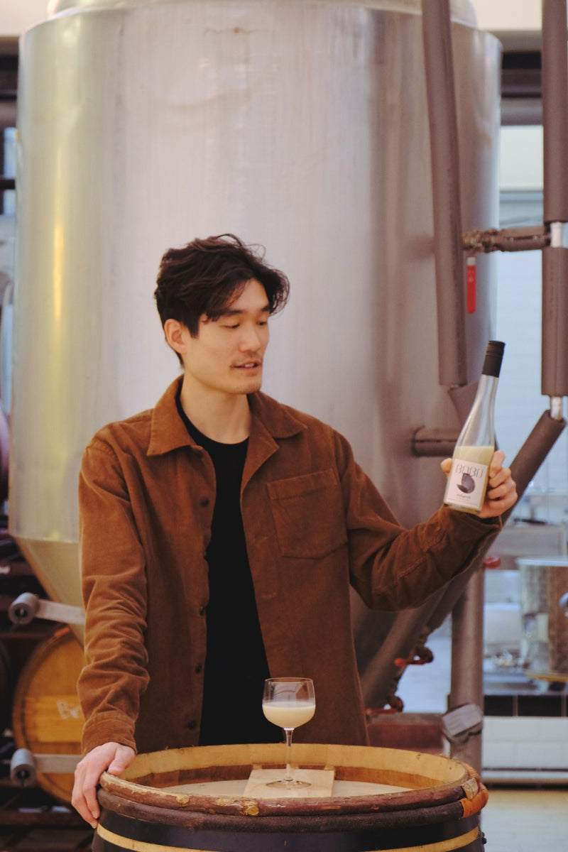 Tim holding up a bottle of Babo Makgeolli and looking at it, with a glass of makgeolli placed in front.